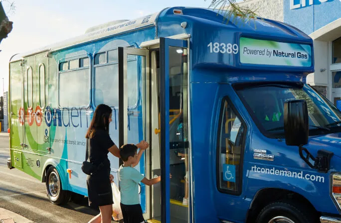 A woman and a child are boarding a blue and green shuttle bus labeled “Powered by Natural Gas” on the side, parked by a sidewalk on a sunny day.