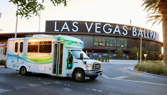 A small shuttle bus with green and blue accents drives past Las Vegas Ballpark, which has large white letters on its exterior. Palm trees and landscaping are visible in the foreground.