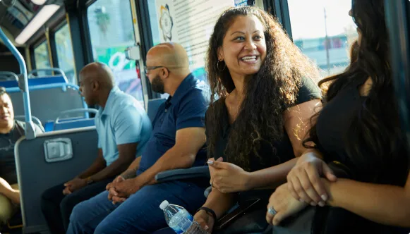 A woman smiles while sitting on a bus, holding a water bottle. Other passengers are seated around her, engaged in conversation or looking out the window. Sunlight streams through the windows, creating a warm atmosphere.