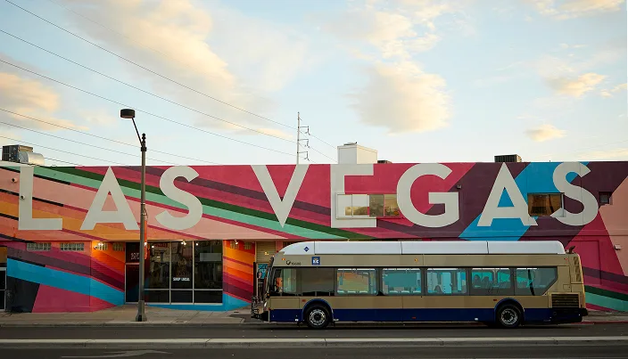 A public bus is parked in front of a colorful mural with large letters spelling “LAS VEGAS” on the side of a building under a partly cloudy sky.