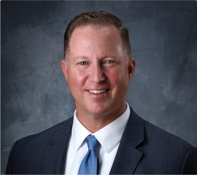 A man in a suit and blue tie smiles at the camera against a gray textured background. He has short, neatly styled hair and a light complexion.