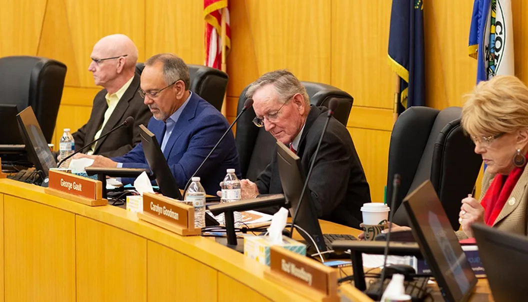Four people sit at a long desk in a formal meeting room with laptops, nameplates, documents, and flags behind them. They appear focused, reading or writing notes during a session.