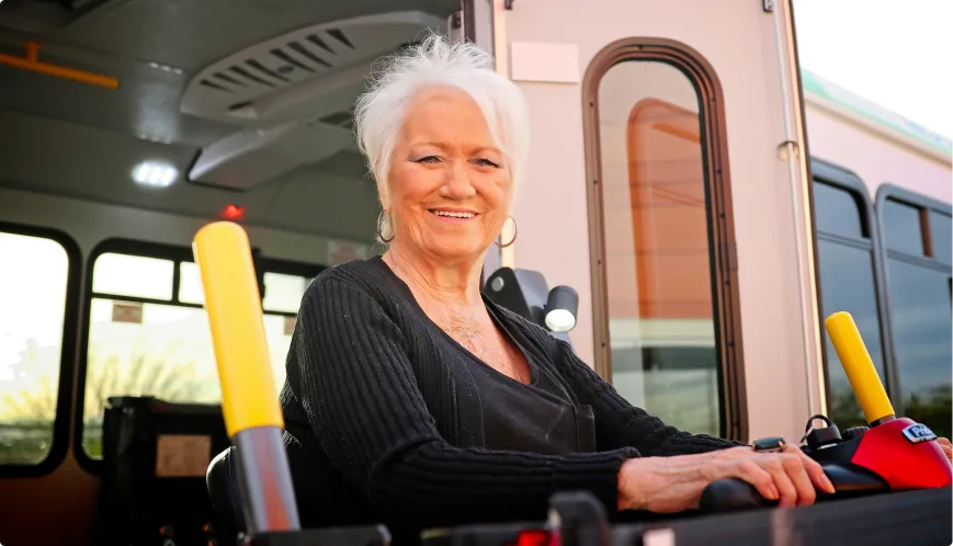 An older woman with white hair smiles while sitting in a wheelchair lift outside a bus, preparing to board. Bright yellow safety bars and the bus entrance are visible behind her.