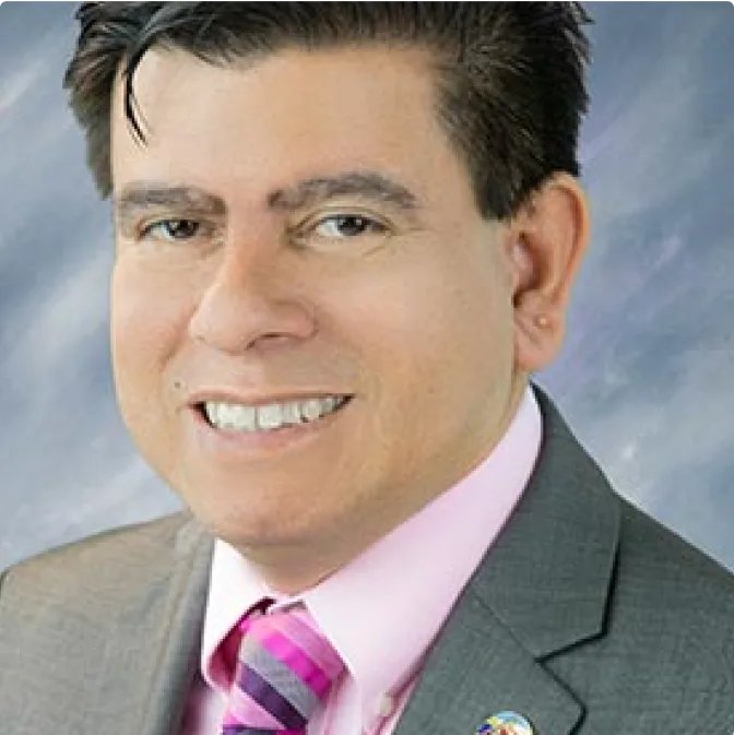 A man with short dark hair wearing a gray suit, pink shirt, and striped pink tie smiles at the camera against a soft, blurred background.