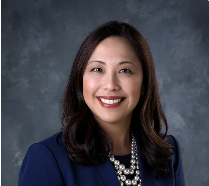 Professional portrait of a woman with long dark hair wearing a navy blazer, white blouse, and layered pearl necklace, smiling in front of a gray textured background.