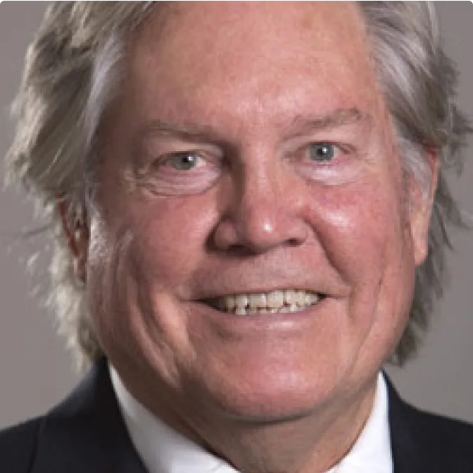 A smiling older man with gray hair wearing a dark suit jacket and white shirt, shown in a close-up portrait against a neutral background.