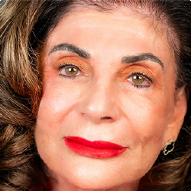Close-up of an older woman with wavy brown hair, bold red lipstick, well-groomed eyebrows, and gold hoop earrings, looking directly at the camera with a neutral expression.