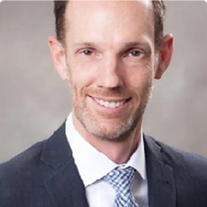 A man with short brown hair and a trimmed beard, wearing a dark suit, white shirt, and patterned tie, smiles at the camera against a plain, light gray background.