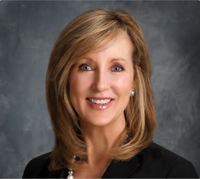 A woman with shoulder-length blonde hair, wearing a black blazer and pearl necklace, smiles in front of a gray background.