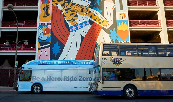 Two buses are parked side by side in front of a colorful mural featuring a tiger and abstract shapes. One bus is labeled Powered by Hydrogen and Be a Hero. Ride Zero, while the other is a double-decker bus.