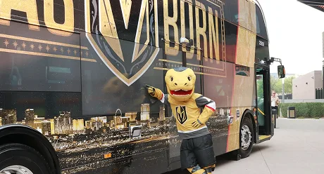 A person in a yellow lizard mascot costume wearing a Vegas Golden Knights jersey poses and points at a large team-themed bus with gold and black graphics.