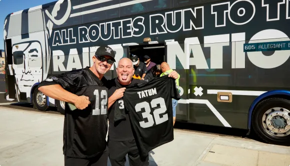 Two smiling men in Raiders jerseys pose in front of a Raiders Nation bus, one holding a Tatum 32 jersey and the other flexing his arm. Other people are seen boarding or exiting the bus in the background.