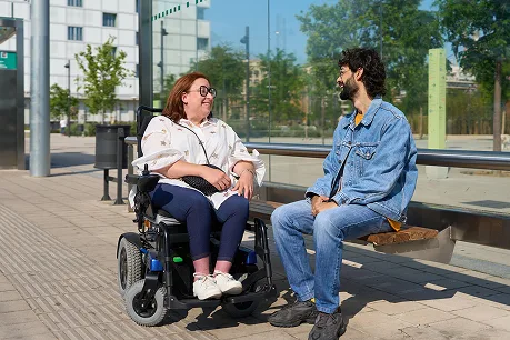 A woman in a wheelchair and a man in a denim jacket are smiling and talking at a sunny outdoor bus stop. Trees and modern buildings are visible in the background.