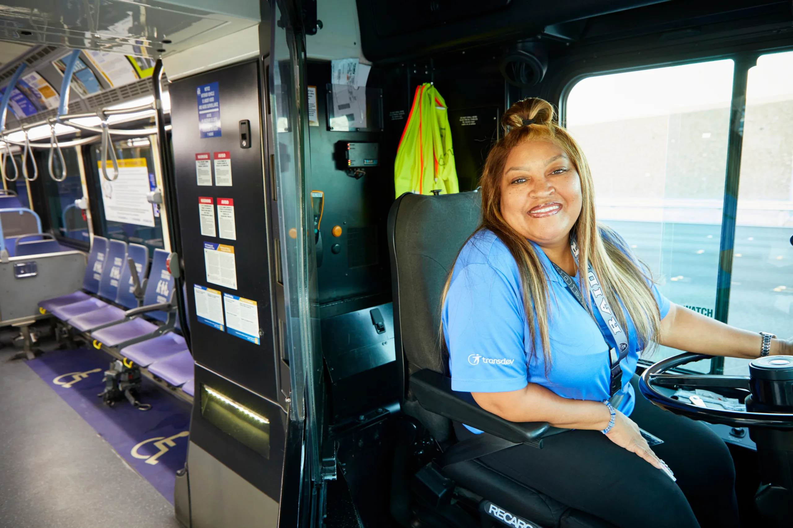 017f6d128471afb044ecf19a388e8216b511b96d A smiling bus driver wearing a blue shirt sits in the driver’s seat of a city bus. The bus interior is visible, with empty purple seats and sunlight coming through the windows.