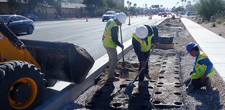 Three construction workers in safety gear are shoveling dirt and working on a road median. A construction vehicle is nearby and traffic cones line the street. The sidewalk and newly built median are visible.
