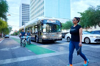 A woman walks across a city street in front of a public transit bus, while two people ride bikes in a bike lane. Tall buildings and trees line the street in the background.