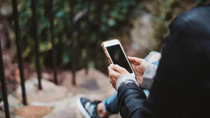 A person wearing a dark jacket sits outdoors on stone steps, holding a smartphone with both hands. The phones screen is dark, and the background is softly blurred with railings and greenery.