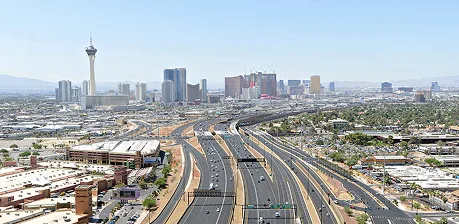 Aerial view of a wide highway with light traffic leading toward the Las Vegas Strip, with tall hotels, casinos, and the Stratosphere Tower visible in the distance under a clear blue sky.