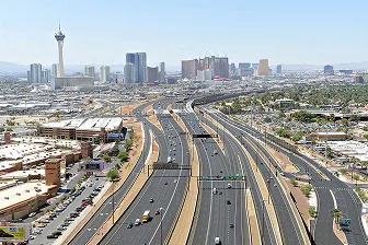 Aerial view of a wide highway with multiple lanes leading toward the Las Vegas skyline, including the Stratosphere tower, with buildings, palm trees, and clear skies in the background.