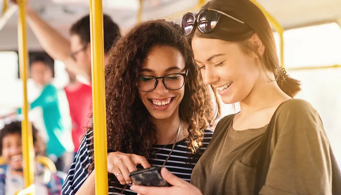 Two young women smiling and looking at a phone together while standing on a bus. Sunlight shines in, and other passengers are visible in the background.