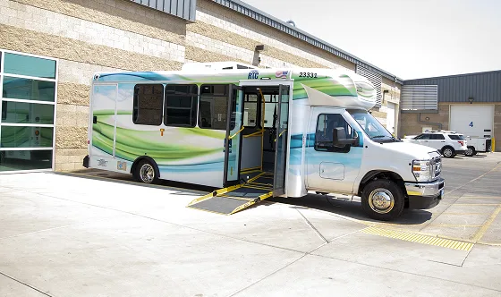 A small city bus with an extended wheelchair ramp is parked outside a building on a sunny day. The bus has a blue, green, and white design and is positioned next to a sidewalk with yellow tactile paving.