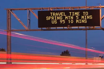 A digital highway sign displays Travel time to Spring Mtn 5 mins, US 95 10 mins above a road with red light streaks from passing vehicles at dusk.