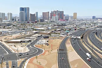 Aerial view of a busy highway interchange with cars and trucks, set against the backdrop of tall modern buildings and hotels in a city skyline, with desert landscaping in the foreground.