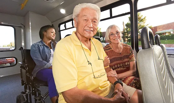Three smiling people sit inside a bus; an elderly man in a yellow shirt sits in front, an elderly woman in patterned dress sits beside him, and a young woman in a wheelchair sits behind them. Sunlight shines through the windows.