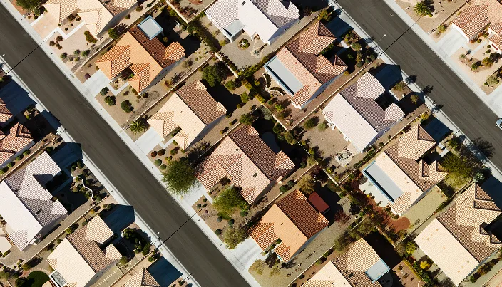 Aerial view of a suburban neighborhood showing rows of houses with tan and orange roofs, separated by streets, driveways, and small yards with minimal greenery.