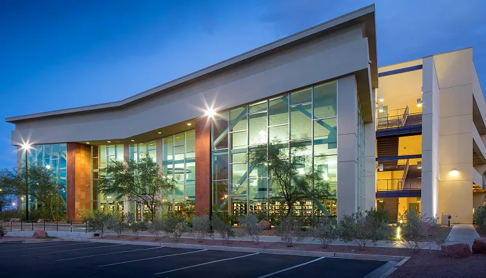A modern, multi-story building with large glass windows is illuminated at dusk. Trees and desert landscaping surround the entrance, and an empty parking lot is visible in the foreground.