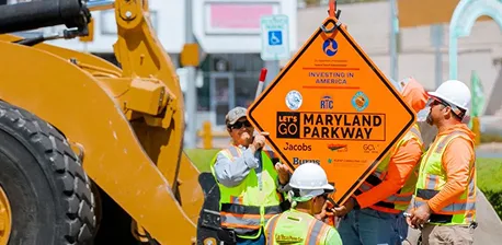 Four construction workers in safety gear hold up an orange diamond-shaped Lets Go Maryland Parkway road sign in front of construction equipment on a sunny day.