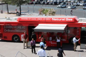 A red bus with Natural Gas written on top is parked outdoors. Several people stand near the bus, engaging at an information booth under an awning. Cars and trees are visible in the background.