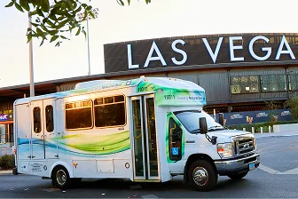 A white and green shuttle bus is parked in front of a building with a large LAS VEGAS sign, with some trees and street elements visible in the foreground.