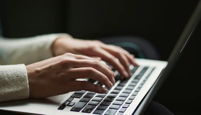 A person wearing a light-colored long sleeve shirt types on a laptop keyboard, with their hands in focus against a dark background.