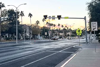 A wide street with cars driving in the distance, palm trees along the sides, a pedestrian crossing sign, traffic lights, and a Stop Here on Red sign by the sidewalk at sunset.