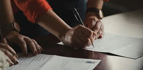 A person wearing a watch and an orange sleeve is signing a document with a pen on a desk, with another persons hand and additional paperwork visible nearby.