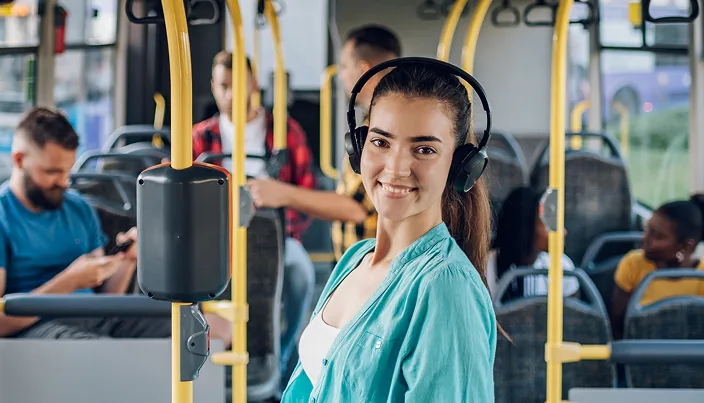A young woman wearing headphones smiles at the camera while standing on a city bus. Other passengers are seated in the background, some looking at their phones or talking.