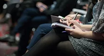 People sitting in rows, taking notes with pens and notebooks during what appears to be a meeting, lecture, or conference. The focus is on their hands and notebooks, with the background slightly blurred.
