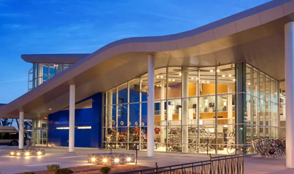 A modern building with large glass windows, white columns, and a wavy roof is lit up at dusk. Bicycles are visible inside through the glass, and a bike rack sits outside.