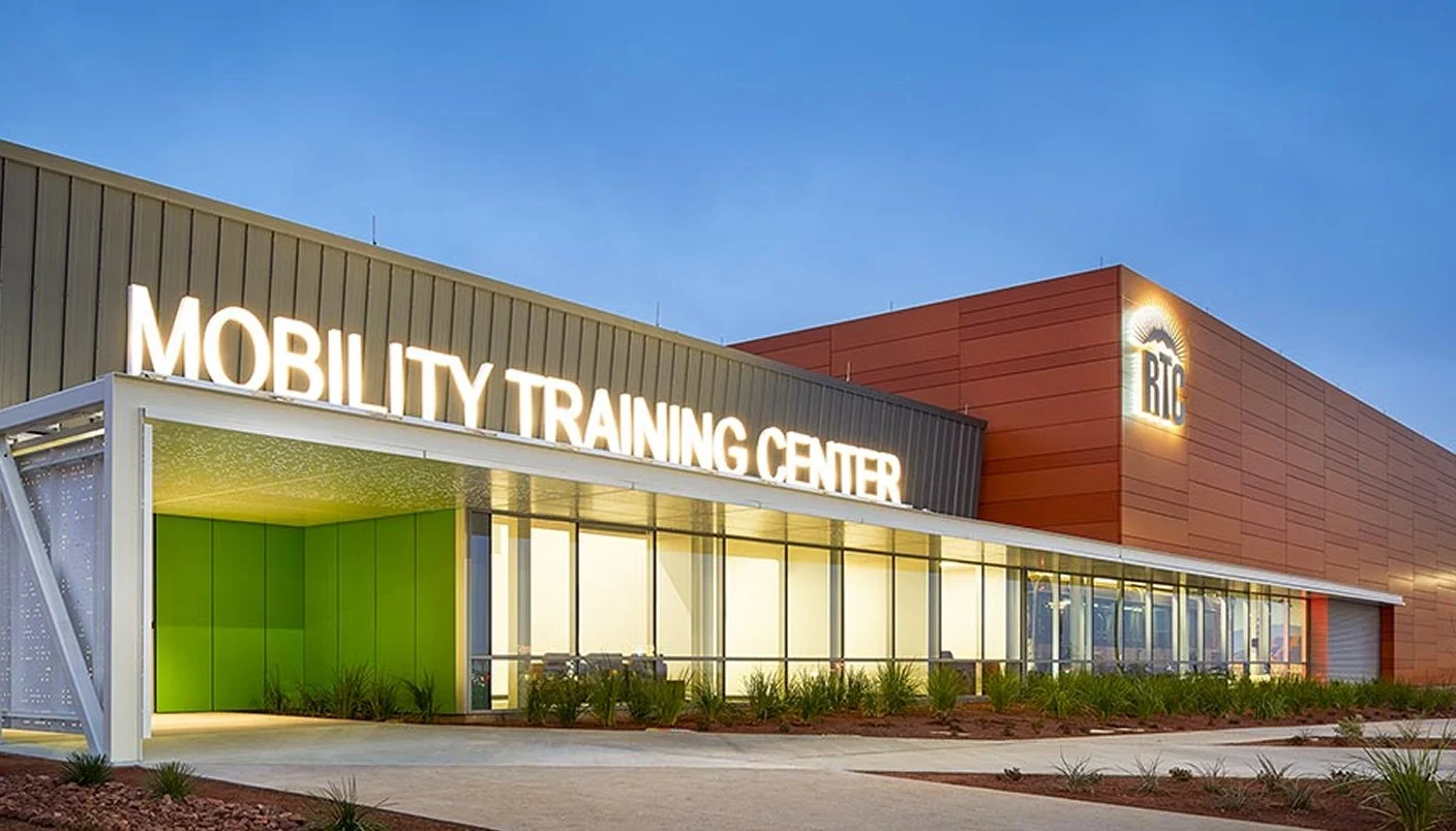 A modern building with large illuminated letters reading Mobility Training Center above the entrance, featuring glass walls, a covered walkway, and landscaping in the foreground.