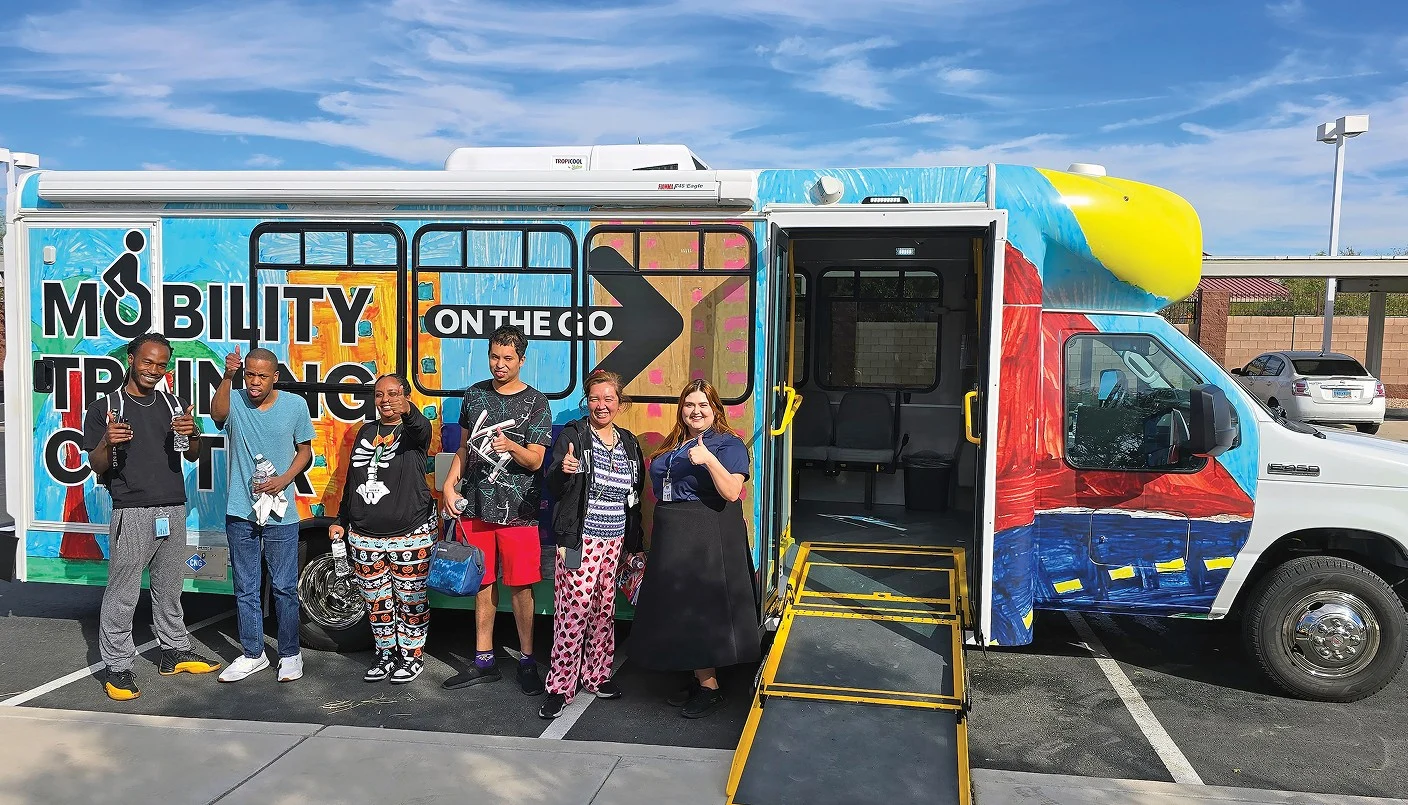 Six people stand and smile in front of a colorful Mobility Training Center bus with a wheelchair ramp extended. The group appears happy, holding snacks or drinks, on a sunny day in a parking lot.