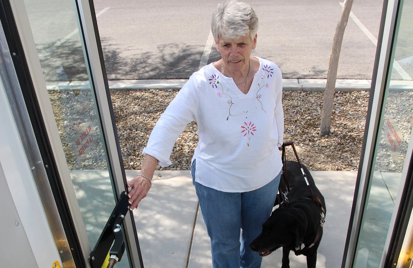 An older woman with gray hair, wearing a white embroidered shirt and blue jeans, enters through a glass door while holding a handle. She is accompanied by a black guide dog in a harness.