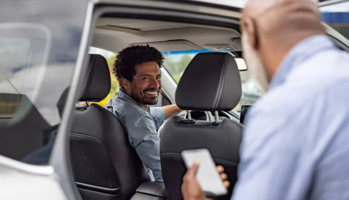 A smiling man in the driver’s seat of a car looks back at a passenger, who is holding a smartphone while sitting in the back seat. The photo is taken from outside the open car door.