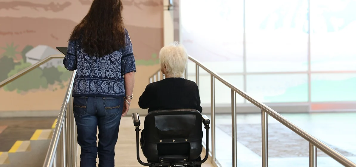 A woman walks beside an older person in a wheelchair as they ascend a ramp indoors, with handrails on both sides and a brightly lit background.
