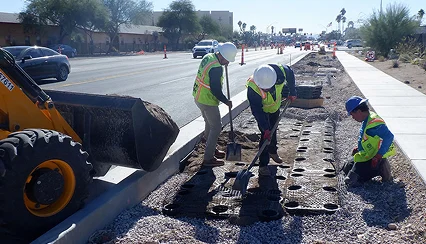 Construction workers in safety vests and helmets install modular paving panels on a sidewalk median, using shovels to spread gravel, next to a busy road with traffic cones and construction equipment visible.