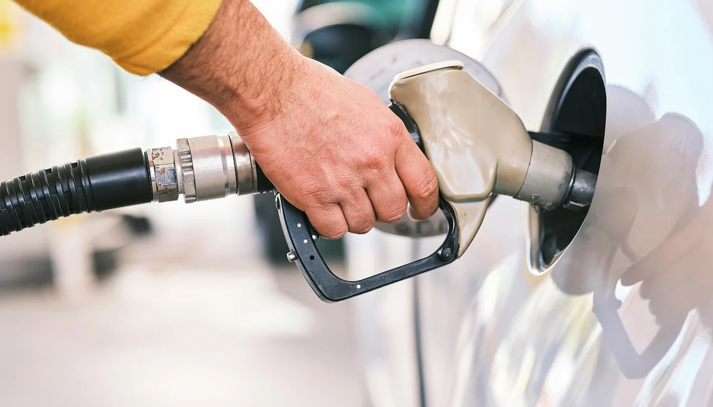 A close-up of a person’s hand holding a fuel nozzle, pumping gasoline into a car at a gas station. The car’s fuel tank door is open, and the nozzle is inserted.