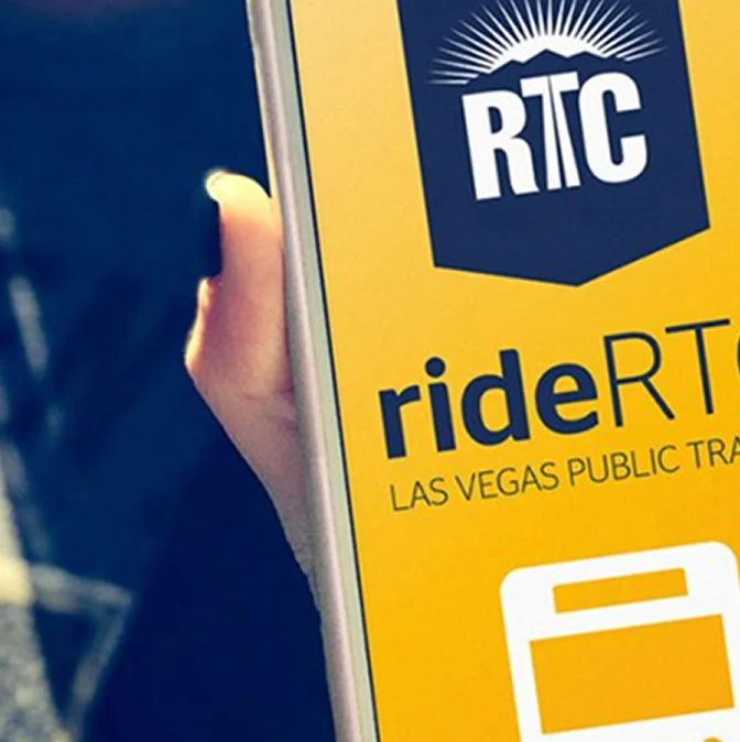 A hand holding a smartphone displaying the rideRTC app, with the RTC logo and the text rideRTC LAS VEGAS PUBLIC TRA visible on a yellow background.