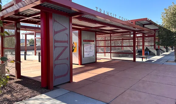 A modern outdoor transit center with red metal framework, shaded seating areas, and a few people sitting on benches. A route map and transit information are displayed on a central board. Trees provide shade nearby.