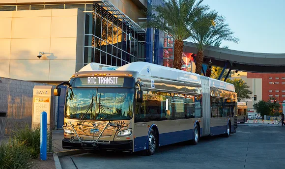 A modern RTC Transit bus is parked at a station surrounded by palm trees and contemporary buildings under clear daylight.