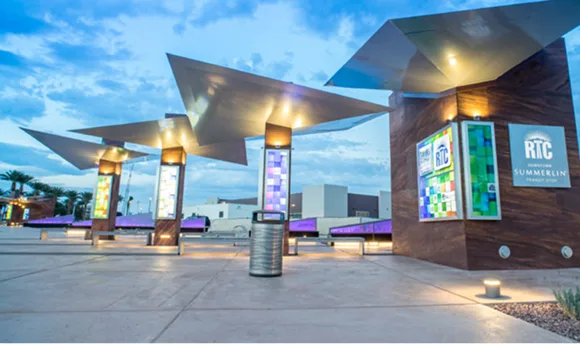 Modern bus stop with angular metal canopies, colorful panel displays, and RTC Summerlin signage. Purple-lit benches and a trash bin are visible, with buildings and palm trees in the background under a cloudy sky.